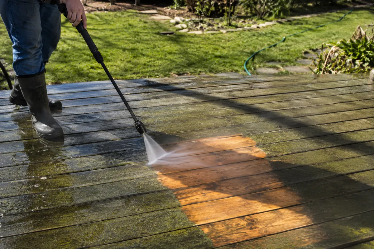 A homeowner pressure-washes algae off a wooden deck with mist catching the sunlight
